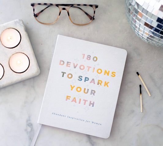 Book titled '180 Devotions to Spark Your Faith' on a marble surface with candles and glasses.