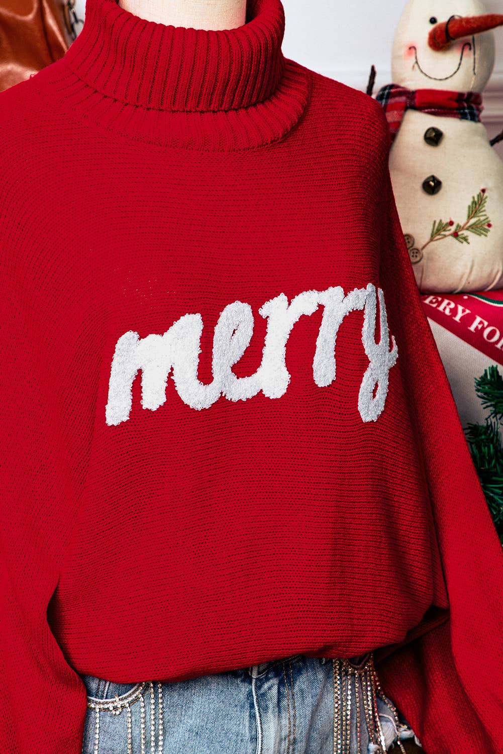 Red sweater with 'merry' text worn by a person, with a snowman decoration in the background.