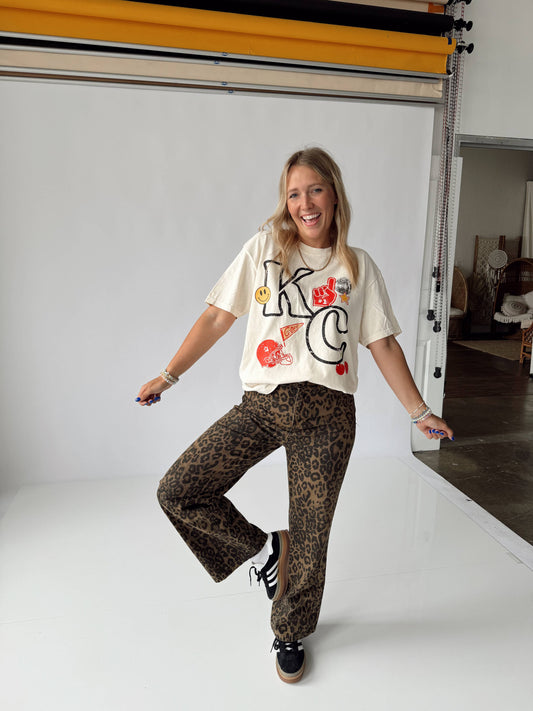 A person wearing a white KC themed t-shirt and animal patterned pants, standing in a room with a white background.