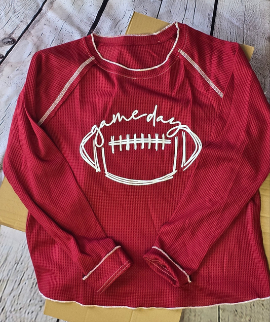 Red long sleeve knit top with a football, gameday in cursive on the front displayed on a barnwood background with over a shopping box