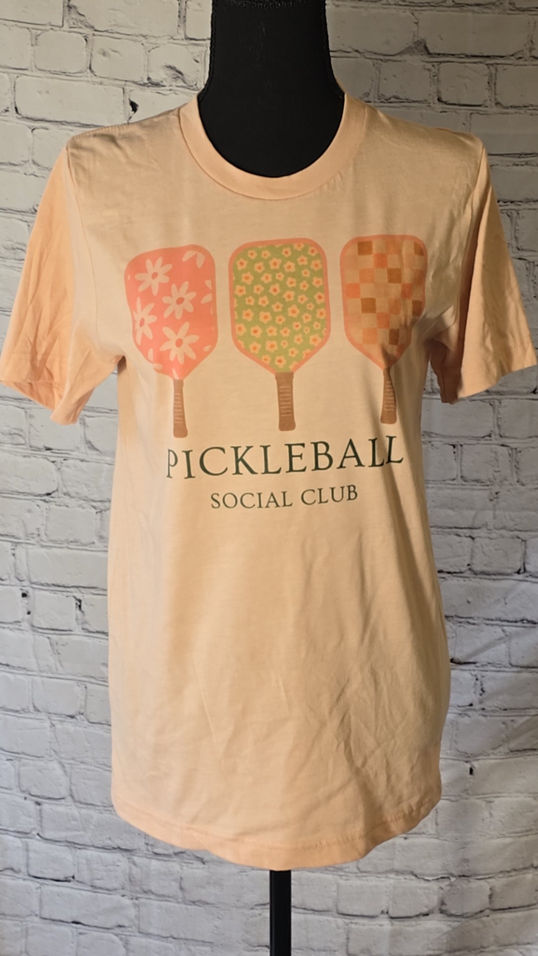 peach t-shirt with 'PICKLEBALL SOCIAL CLUB' text and a graphic of three pickleball paddles in pink, green, and yellow colors with floral and checkered patterns.