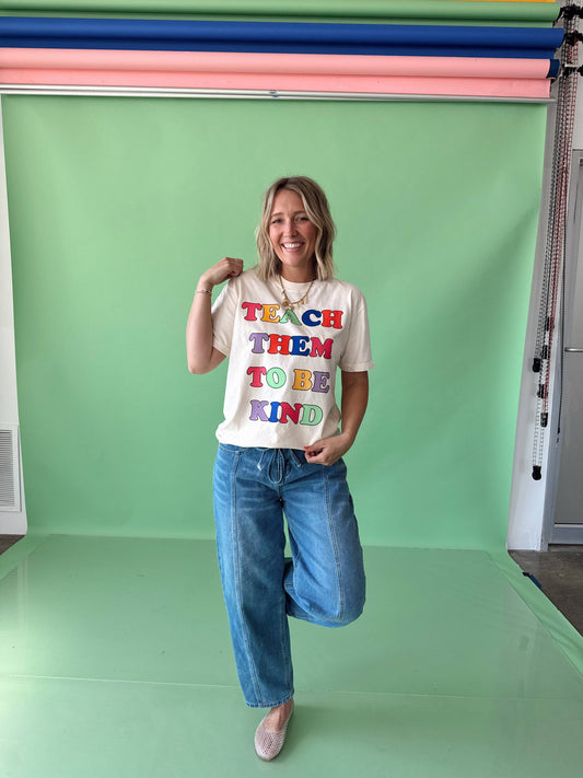 A person wearing a white t-shirt with the text 'TEACH THEM TO BE KIND' in colorful letters, paired with blue jeans and beige shoes.