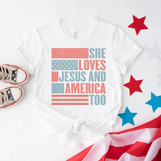 White t-shirt with 'She Loves Jesus and America Too' text, surrounded by American flag and star decorations on a white background.