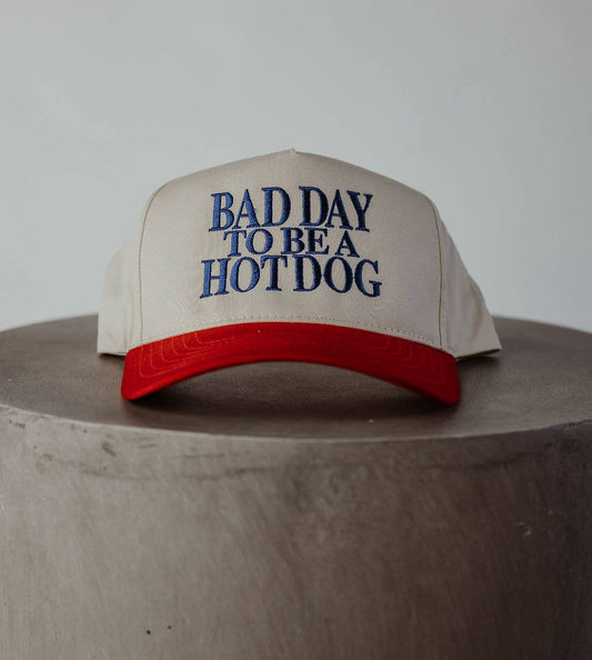 Beige cap with red brim and blue text 'Bad Day to be a Hotdog' on a wooden surface.