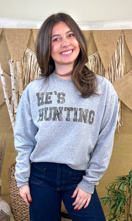 Person wearing a gray sweatshirt with 'HE'S HUNTING' text in front of a decorative wall.