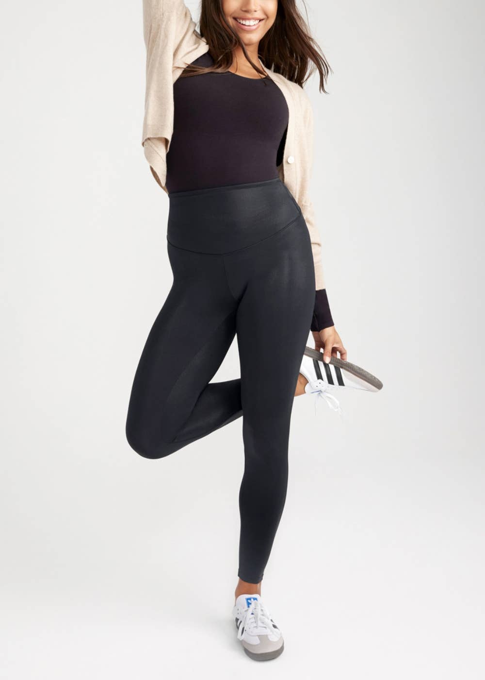 Woman standing with one leg bent bent behind the other on a solid background wearing leggings, a black top and a tan jacket