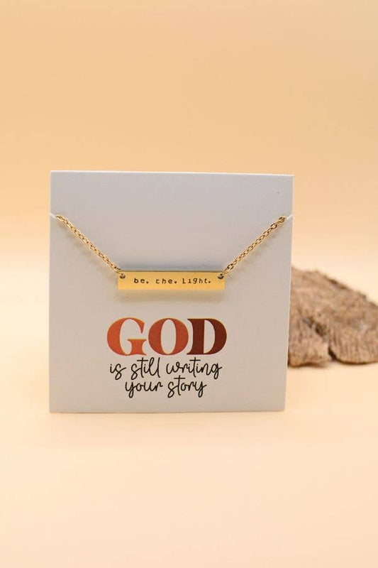 Gold necklace on a card with 'God is still writing your story' text on a beige background