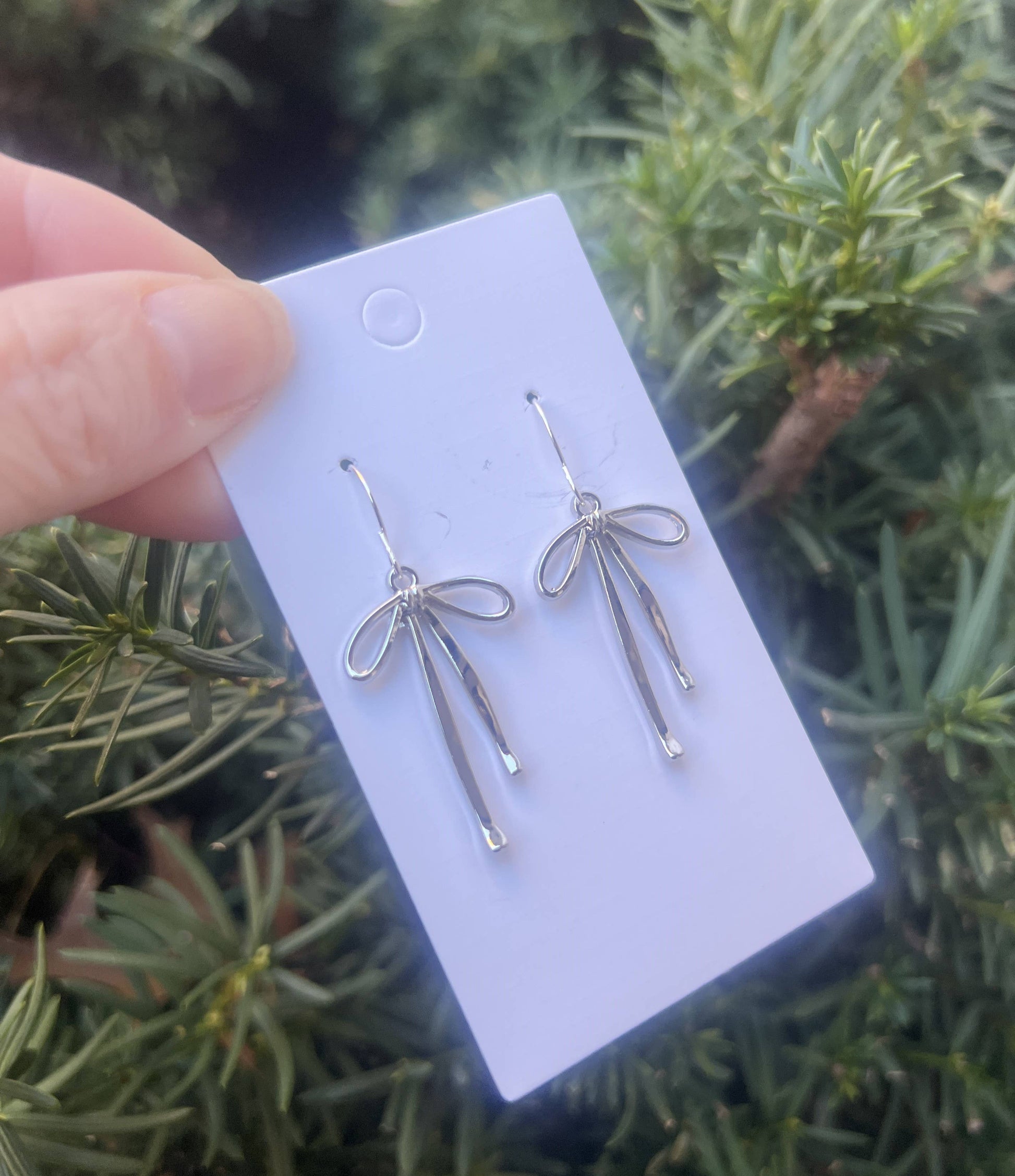 Silver bow-shaped earrings on a white card held against a green foliage background