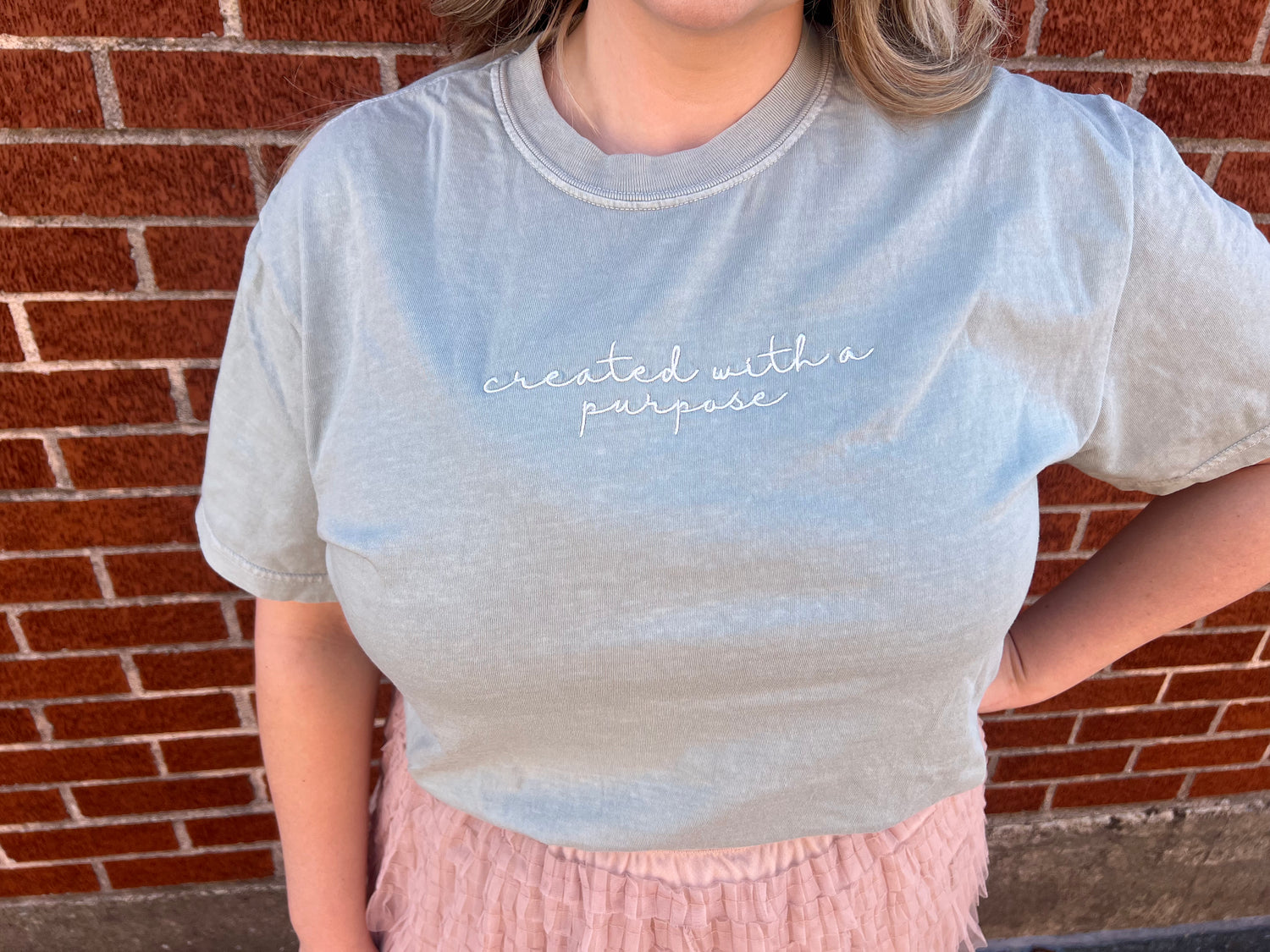 A person wearing a sage colored t-shirt with the phrase 'created with a purpose' embroidered on the front.