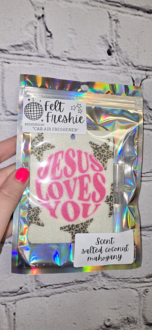 A hand holding a car freshener pack with 'JESUS LOVES YOU' text visible on the front. The pack is predominantly white with a pattern and the text is in pink.